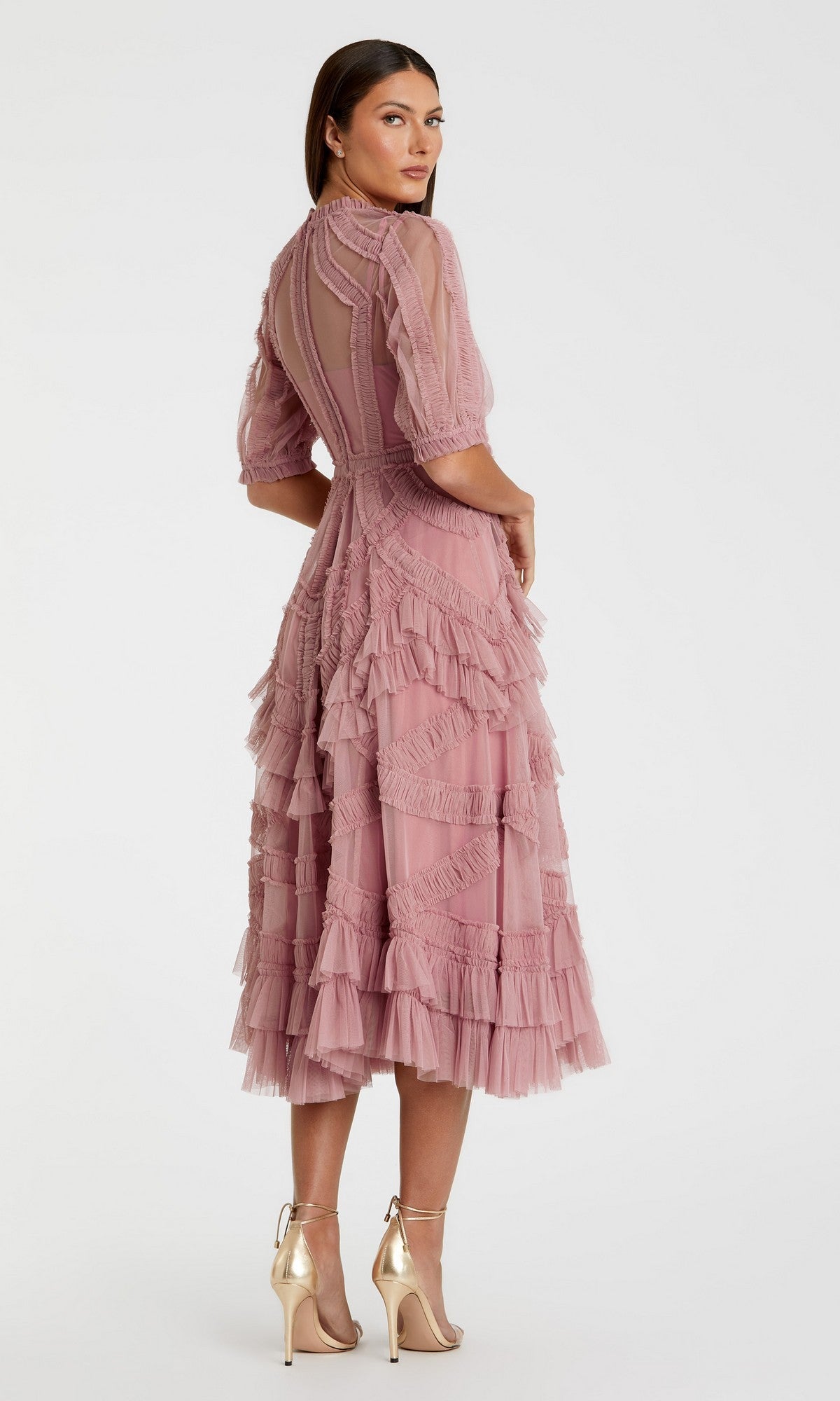 A woman models the Mac Duggal 8058, a mauve ruffled midi wedding-guest dress with sheer sleeves and gold heels, standing against a plain white background.